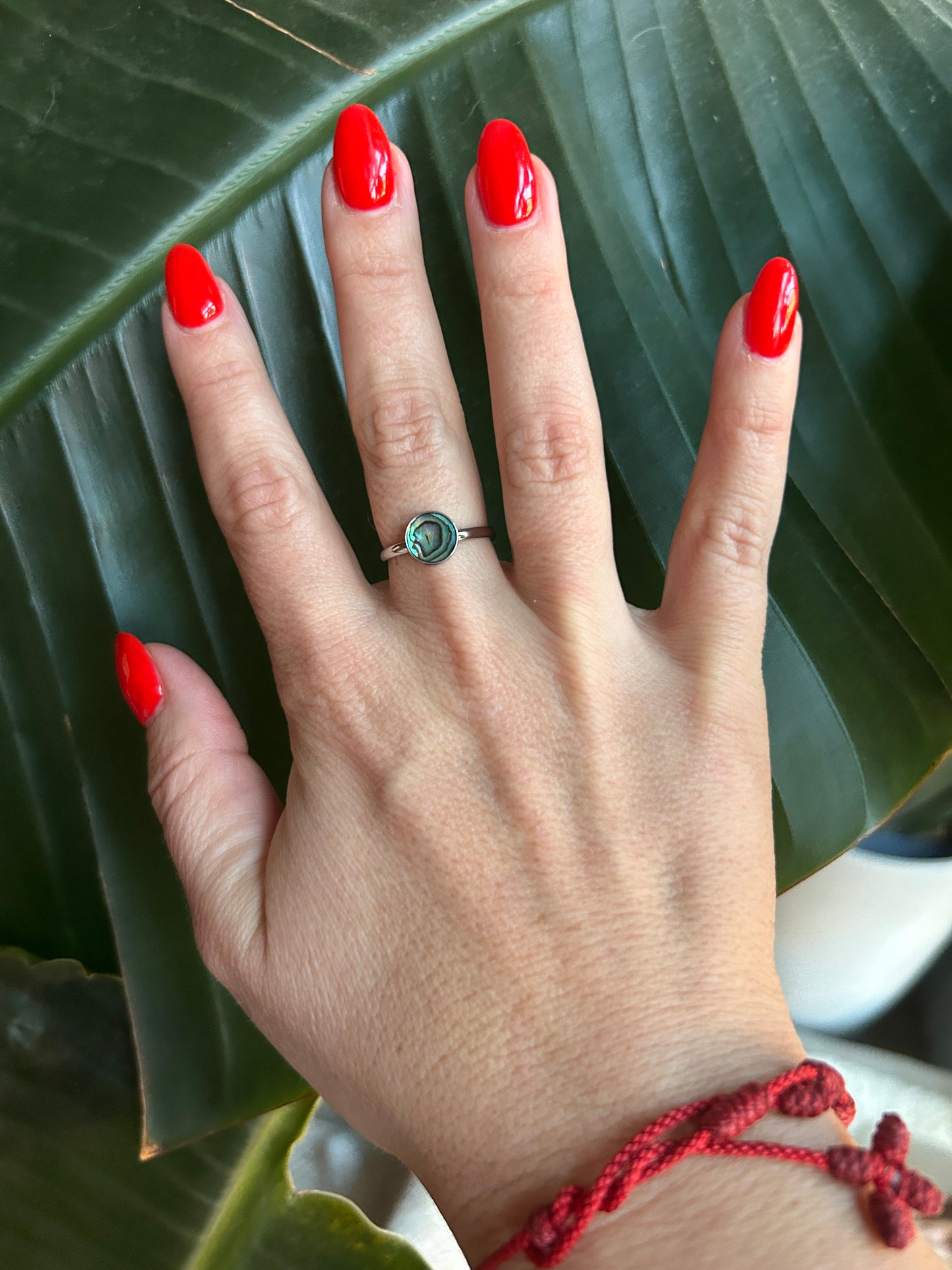 Hand with red nail polish and an abalone ring against a green leaf background
